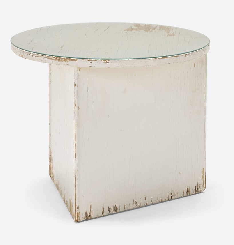 a triangular base table with an offset circular top is made of douglas fir plywood, but is also 90 years old, and the white paint all over it looks like it. a glass top prevents easy access to the storage area inside. being sold by la modern in 2025, originally from rudolph schindler's walker house in silver lake.