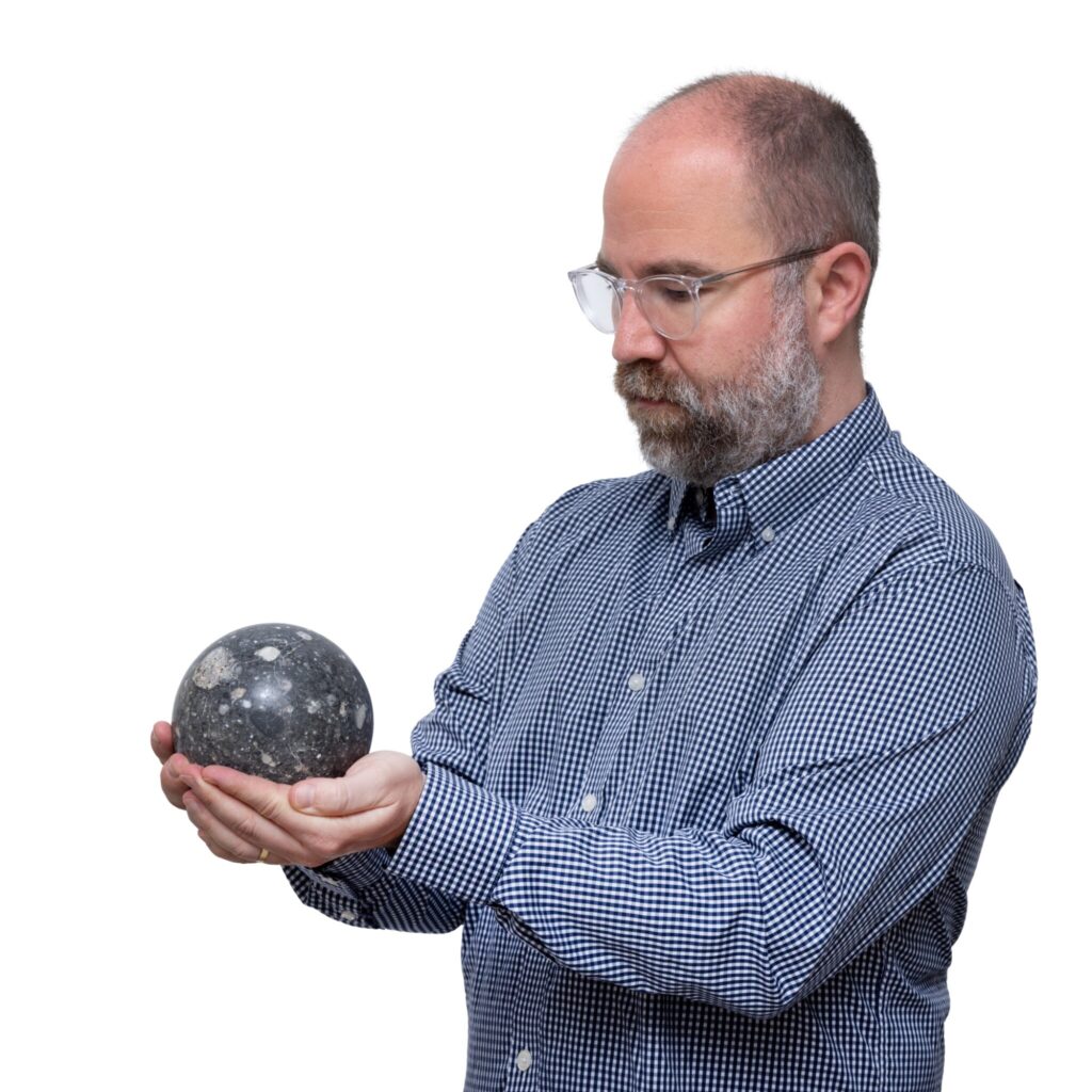 a middle aged white guy with a close cut balding head and salt & pepper beard and glasses wearing a blue checked dress shirt is holding a grapefruit-sized sphere made from a lunar meteorite, which he is selling in his natural history auction at sotheby's new york in july 2025. the sphere is dark grey, mottled with lighter grey spots.