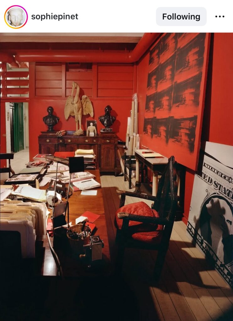 screenshot of sophie pinet's instagram post of a very cluttered office in a loft space full of dark medieval furniture, warhol bric a brac, and a big ass electric chair painting propped on a console behind the desk. it's red, almost exactly the same red as all the walls and ductwork. fred hughes's office in warhol's last factory