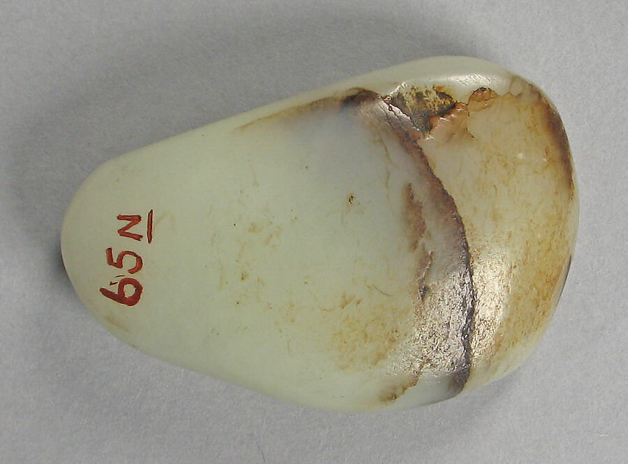 a color photo of a single jade pebble, about 2 inches long, and shaped a bit like a guitar pick, mostly with the ivory color of a human tooth, except for one side which is the browner color of a human tooth of someone who smokes like three packs a day, with the catalogue number 65 N in red paint on one end, from the metropolitan museum