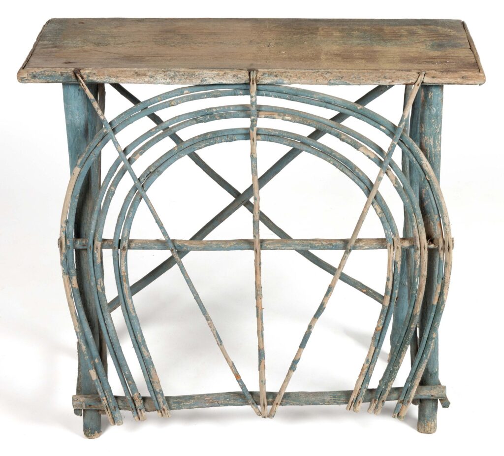 an appalachian twig side table whose main feature is three long arched twigs on the front held by three straight sticks coming to a point at the bottom, and a stabilizing x of twigs on the back. the lower twigs are all painted in pale blue, a long time ago. selling in august 2025 at jeffrey s evans in shenandoah somewhere