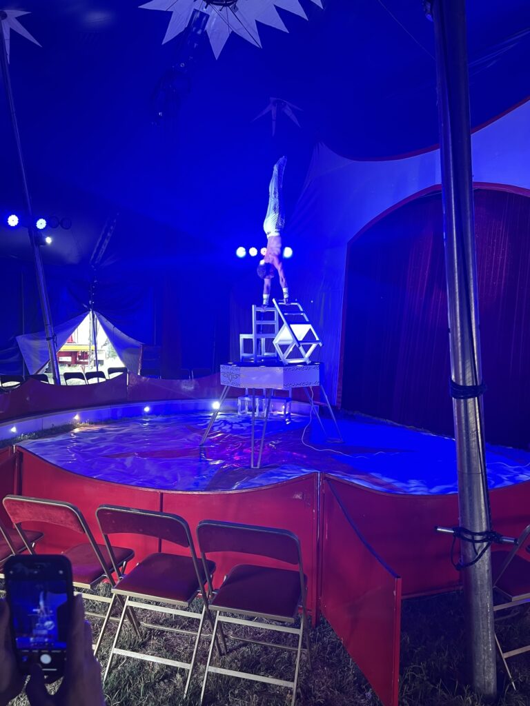 in the center of the small ring of what looks to be an empty traveling circus, except it's really not, it's just that only like eight people paid the extra five euros for the front row seats, a white guy with no shirt and long white pants does a handstand on a pair of white chairs atop a hexagonal raised platform ringed with spotlights. the whole scene is inside a dark blue circus tent, and a triangular side door slash vent reveals the brightness of the still hot af evening in provence.
