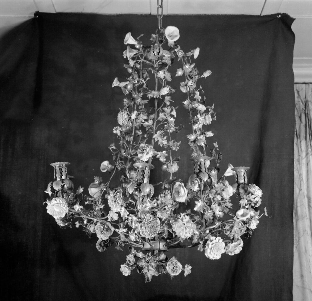 an 18th century rococo chandelier for nine candles is covered with porcelain flowers of various types in this 1974 photo documenting its accession into the metropolitan museum's collection. the photo, taken in the donor's home, is notable to me, anyway, for having a dark background fabric just tacked to the ceiling behind it in a way that exposes the wall and the whole situation