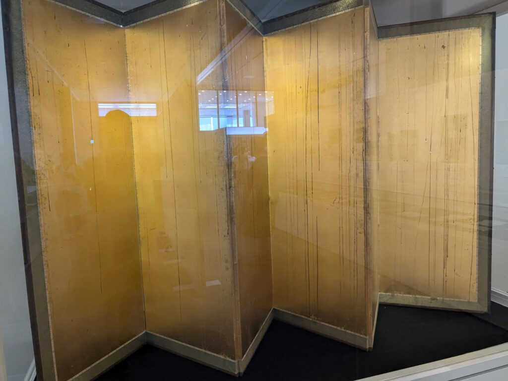 a six panel gold leaf folding screen, commonly known in japan as a kinbyoubu, is streaked with long vertical traces of sooty black, left by black rain falling through the damaged roof of a house in hiroshima after the united states dropped an atomic bomb on the city in 1945. the screen here sits within a glass vitrine at the peace memorial museum, and the exterior windows of the museum reflect in the vitrine glass. this photo was taken by bryan hilley in october 2024 and posted to bluesky in august 2025