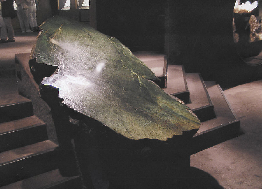 a color photo of a 7-ft long jade boulder, deep green, with a curved, sloping surface, mounted between two low flights of stairs covered in dark chocolate carpet, the lost conversation pit-style hall of minerals at the american museum of natural history, where this jade boulder has been on loan from the met since 1906.