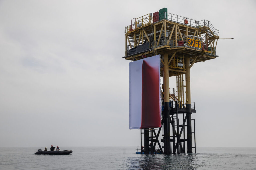 a shell gas drilling platform towers over a zodiac in the north sea. on the side facing the little boat of greenpeace activists hangs a 12m x 8 m white canvas, with a red stain running down the center and right, from an origin point at the top edge, where the activists used a high pressure hose to release 1,000 liters of non-toxic dye to realize anish kapoor's protest artwork idea. via greenpeace