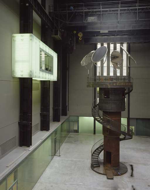 a nine meter tall cylindrical metal tower sits in the vast turbine hall of tate modern, with a glass box window from a gallery on the adjacent left wall. the tower by louise bourgeois is wrapped by a spiral staircase and topped by a platform on which are mounted four large round mirrors on tall stands, reflecting and magnifying the people on the platform and displaying them to viewers around the hall. via tate