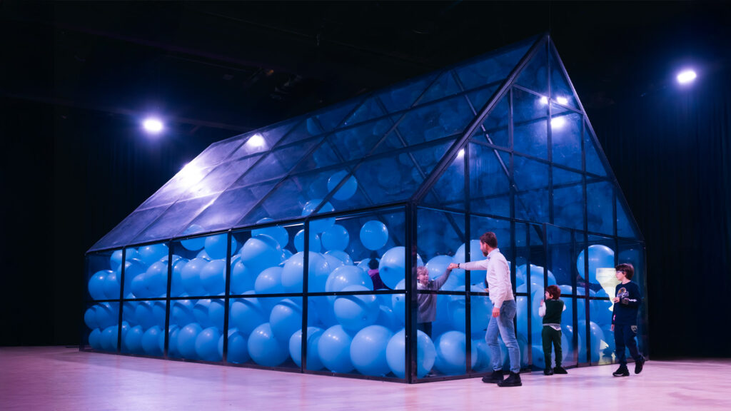 a sadly dimensioned fake greenhouse supposedly half filled with beach ball sized light blue balloons is photographed brightly lit from the outside in an otherwise darkened space. a white dad with two kids on the outside gestures toward another kid trapped inside. from the balloon museum's offensive installation of martin creed's iconic work at the grand palais