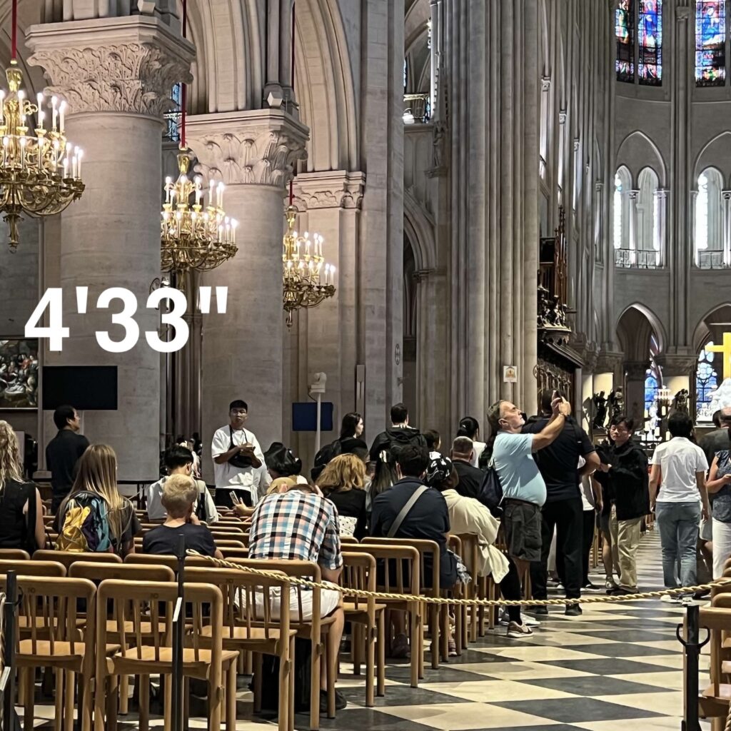 the interior of notre dame cathedral in paris with a row of arched and columns along the left side of a section of wooden chairs in rows, roped off for crowd flow, and about half full of people sitting. in the central aisle with a diamond patterned stone floor a white guy with a gut in a blue polo shirt has his arm raised to take a photo. in the distant background on the right are the apse with tall narrow stained glass windows, taken in aug 2025. also there is a title, 4'33", in white helvetica on the center left of the image, because this is a cover image for a performance of john cage's work now.