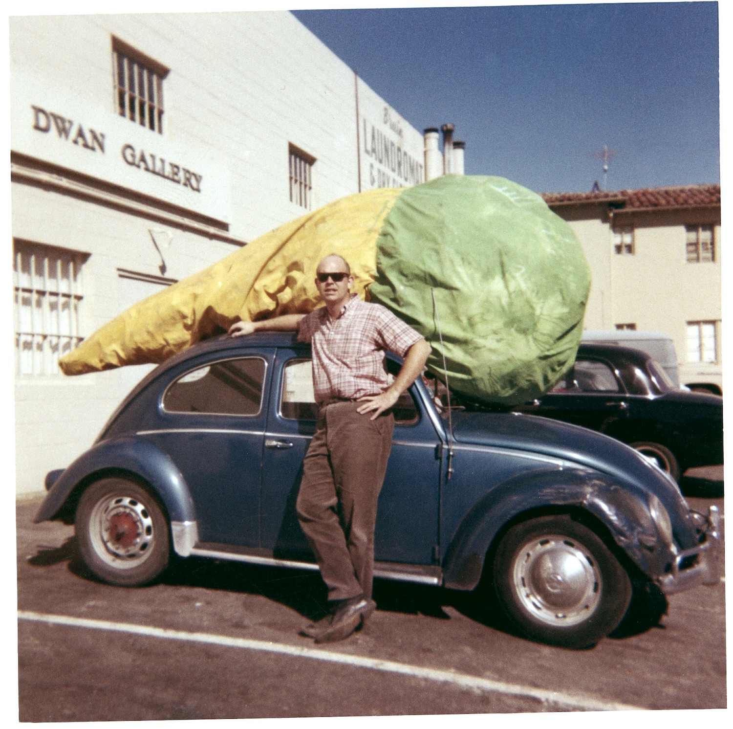 a geeky young claes oldenburg poses for a photo in the parking lot of the virginia dwan gallery in los angeles in 1963. he wears a brown plaid shirt and pants in a forgettable dark color. he rests his arm on the roof of a dark blue vw beetle that has some damage to the front fender. sitting on top of the car is oldenburg's 1962 sculpture, floor cone, an ice cream cone made of roughly painted canvas, green for the ice cream and tan for the cone, all stuffed with, I believe moma conservators said it was stuffed with thousands of cardboard ice cream cups, but it looks lumpy and a bit pathetic in form, but absolutely magical in scale and subject. an 11-ft long masterpiece made even better by being placed on the roof of a similarly scaled car. the buildings are white and inelegant, the sun is bright the sky is cloudless blue image via the artists and moma