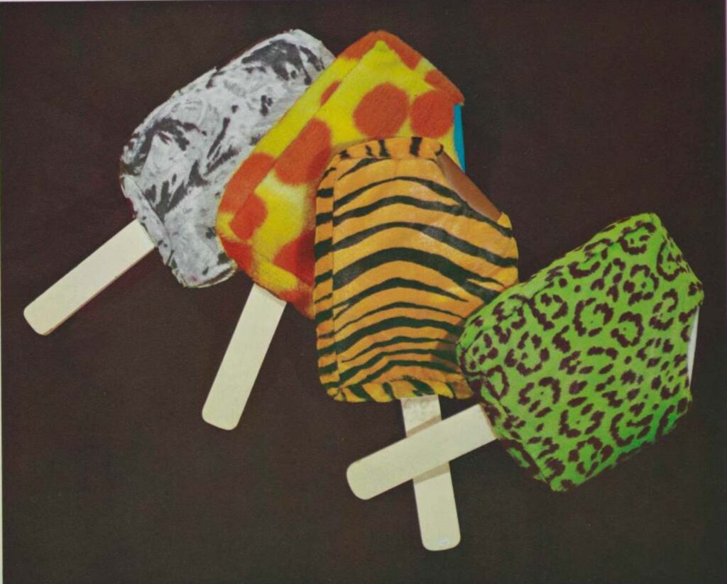 four good humor ice cream bar sculptures made of fake fur fabric in animal skin patterns, but garish colors, against a black background. by claes oldenburg