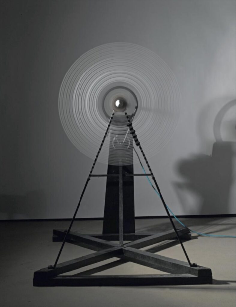 a black painted apparatus on an x-shaped frame holds five glass plates along a spinning axle, with motor at the back. here the timelapse photo depicts the plates spinning so that the stripes painted on them create the optical illusion of concentric circles. the pompidou made this 1979 replica of duchamp's 1920 sculpture, rotary glass plates, under shady museum director pontus hulten, who stole sketches for it from yale university in 1960.
