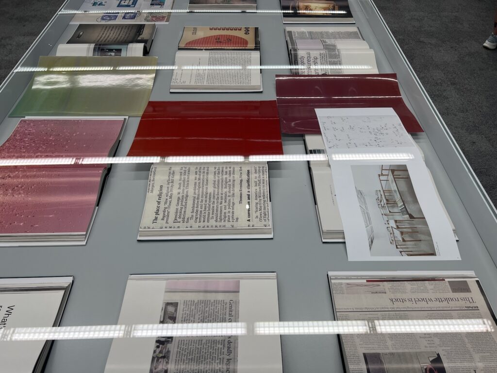 a plexiglass vitrine table at wolfgang tillmans' pompidou exhibition is filled with books opened to pages containing the artist's photos. a red monochrome print at the center is the most prominent of several  prints interlaced within and on top of the books.