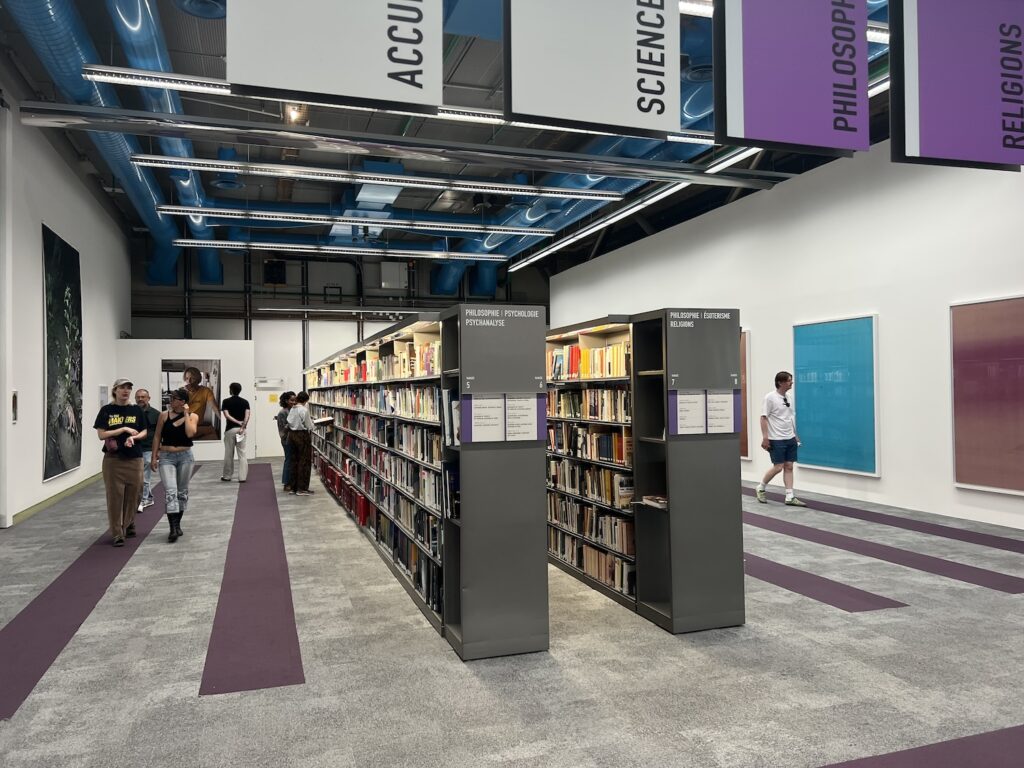 two long grey metal bookcases filled with 12k books, in a space at the pompidou library where purple parallel stripes in the carpet indicate the former presence of more bookshelves. large format photos by wolfgang tillmans hang on walls on either side