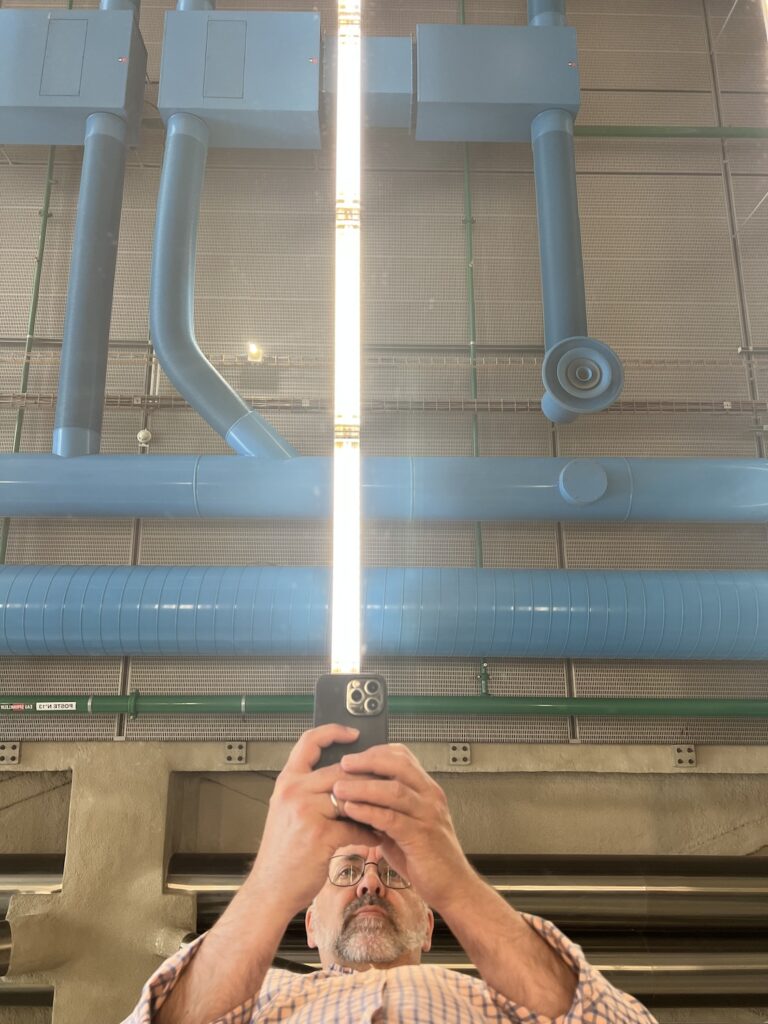 a middleaged white guy in a pale pink-orange checked shirt takes a selfie in the mirrored tabletop surface, with the industrial beams and metalwork of the ceiling reflected. he has lined up the phone in his hands with the bright white fluorescent lamp fixture above to look like a light saber, and so would you. at the wolfgang tillmans at the pompidou
