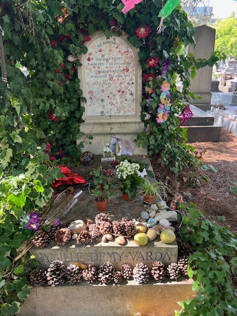 in august 2025 the white marble gravestone of jacques demy and agnes avarda is surrounded by green vines, interwoven with fake flowers, and a string of prayer flags reaches out from the top to the right edge of the image. the gravestone is covered with red lipstick marks and doodles. the grave in front is covered with dead flowers, pine cones, and potatoes, a reference to varda's last good film that didn't involve jr, les glaneurs et la glaneuse.