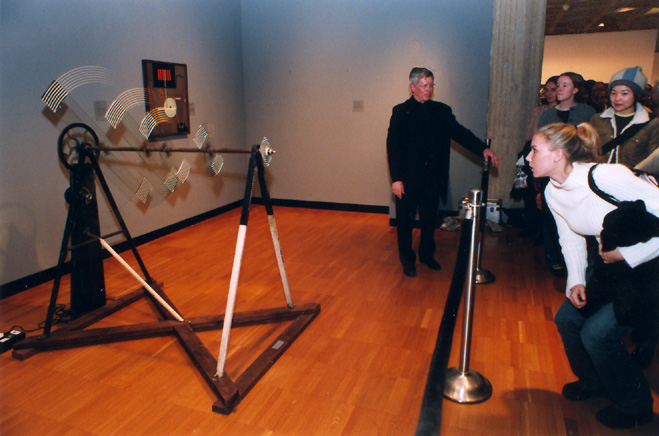 in a warmly lit gallery at yale, a white woman behind a stanchion with her hair pulled back like a soccer player leans down to align her vision with the axle of marcel duchamp's sculpture where five spinning glass plates create the optical illusion of concentric circles. the x-based apparatus has a motor at back. the axle is supported at eye level for a shorter person. duchamp made this in 1920, and katherine dreier donated it to yale in 1941. it is rarely turned on, though, and perhaps the man in front of the stanchion in this photo, wearing an all dark suit, is one of the yale engineering professors who restored the sculpture in 1999. a crowd in the background is perhaps waiting for this white lady to finish so they can take a turn.